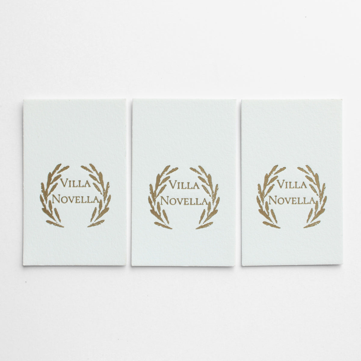 Three identical rectangular white business cards arranged side by side on a white background, each printed in letterpress with the words 'Villa Novella' inside a gold laurel wreath.