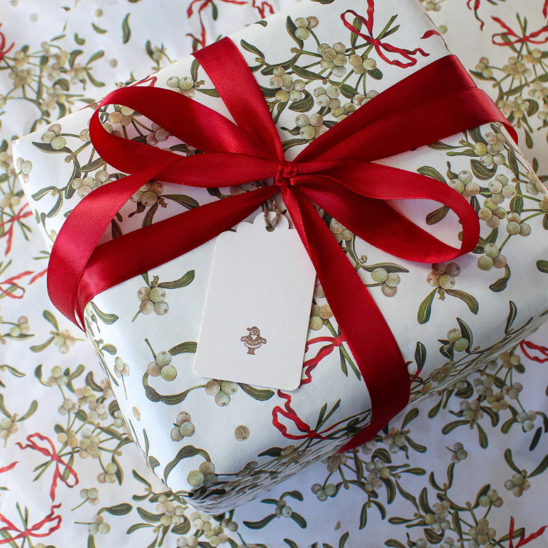 A square gift box wrapped in white holiday paper with a green and beige mistletoe print, tied with a large red satin ribbon and bow, with a white gift tag attached featuring an illustration of father christmas printed in letterpress.