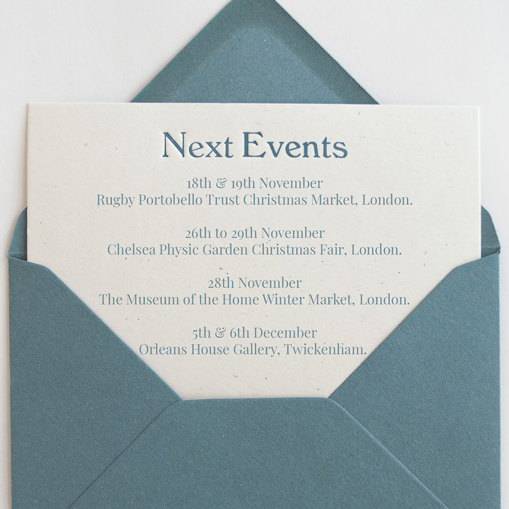 A close-up of a printed invitation card tucked inside a blue envelope. The card reads 'Next Events' with several event dates and locations listed below in blue text. 18th & 19th November is Rugby Portobello Trust Christmas Market, London. 26th to 29th November is Chelsea Physic Garden Christmas Fair, London. 28th November is The Museum of the Home Winter Market, London. 5th & 6th December is Orleans House Gallery, Twikenham.