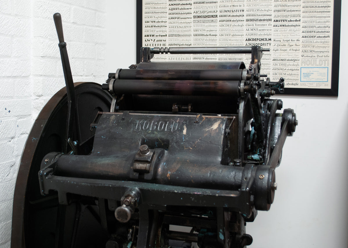 An antique black metal printing press with a large circular wheel on the left, photographed in the corner of a studio with a framed typography poster on the wall behinf the printing press.